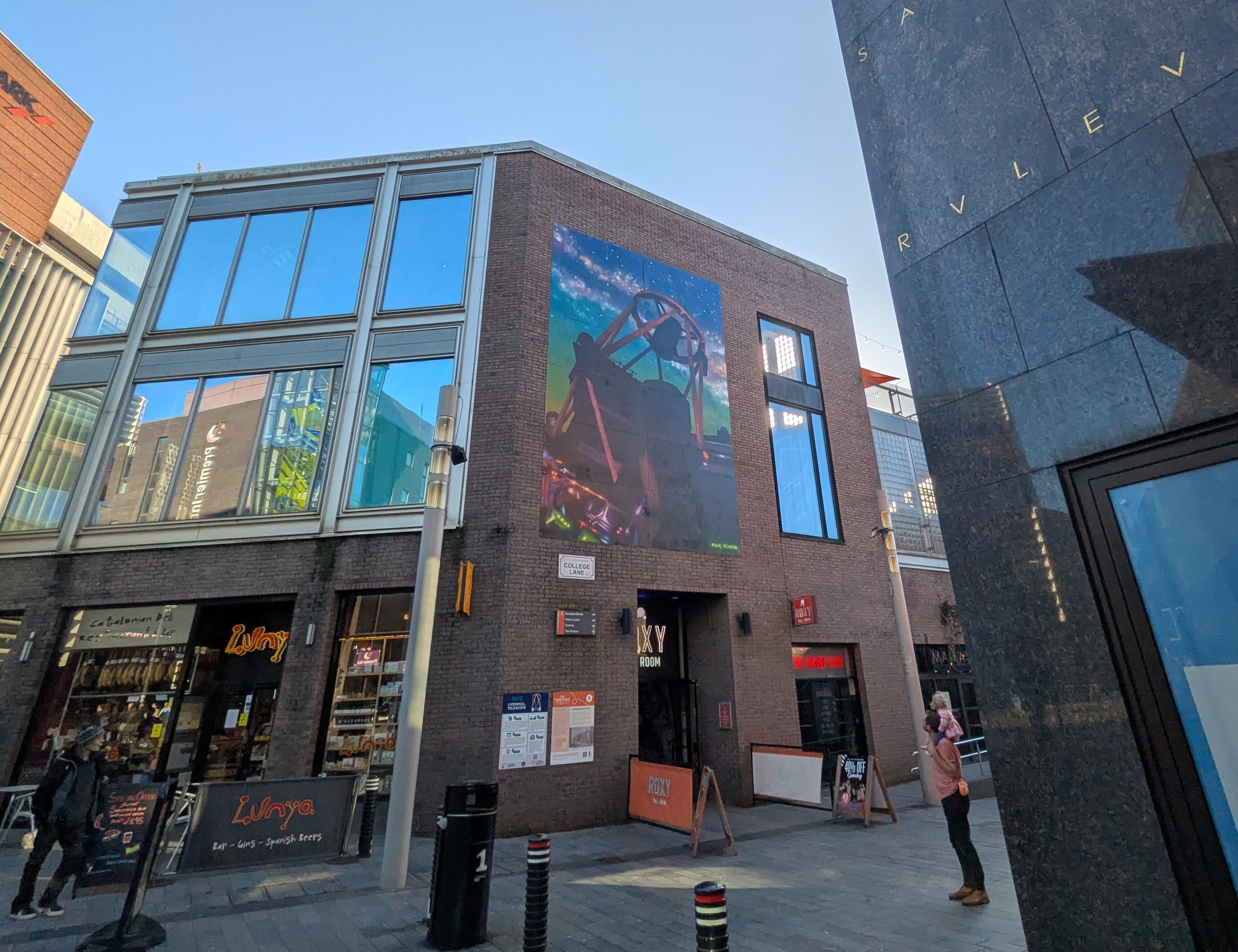 A member of the public carries a child on their shoulders and stops to look up at the LT mural on the side of a building in Liverpool One shopping centre